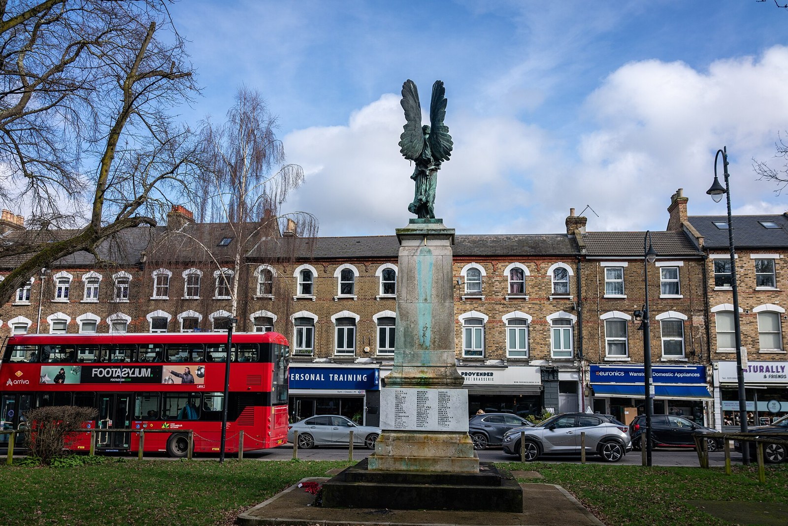 Wanstead war memorial on the green opposite the High Street, with shops and a red bus behind