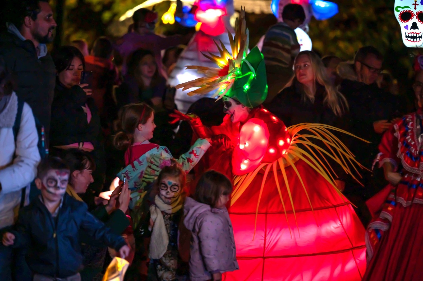 A glowing red lantern puppet towers over children with painted skull faces at a Festival of the Dead parade