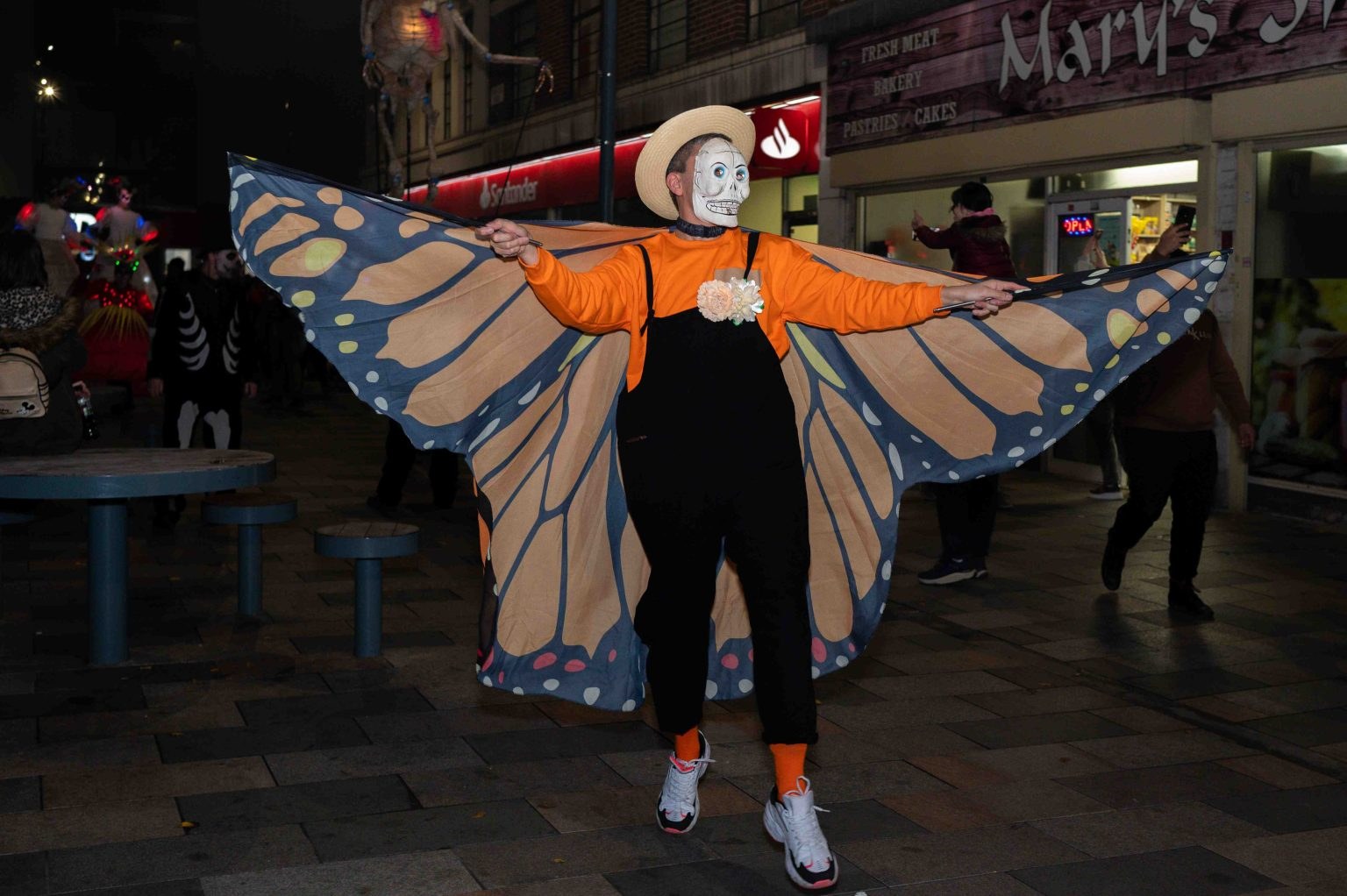 A performer in a skull mask and monarch butterfly wings with arms outstretched in the street at night